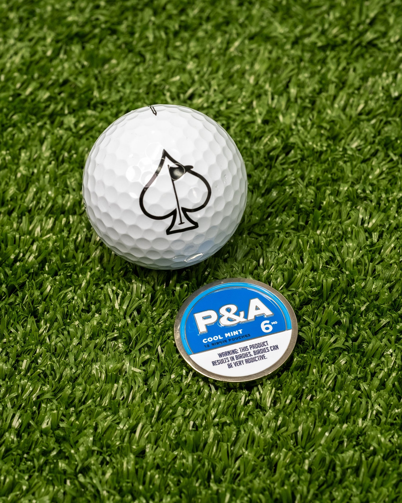 A Pins and Aces 6 Milly - Cool Mint Ball Marker in a round blue tin sits on green turf beside a white golf ball with a black spade symbol. The enamel-painted marker adds style to your game.