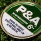 A close-up of a round green and white Pins and Aces 6 Milly - Wintergreen Ball Marker sits on artificial grass next to an enamel golf marker, labeled: "This product results in birdies. Birdies can be very addictive.