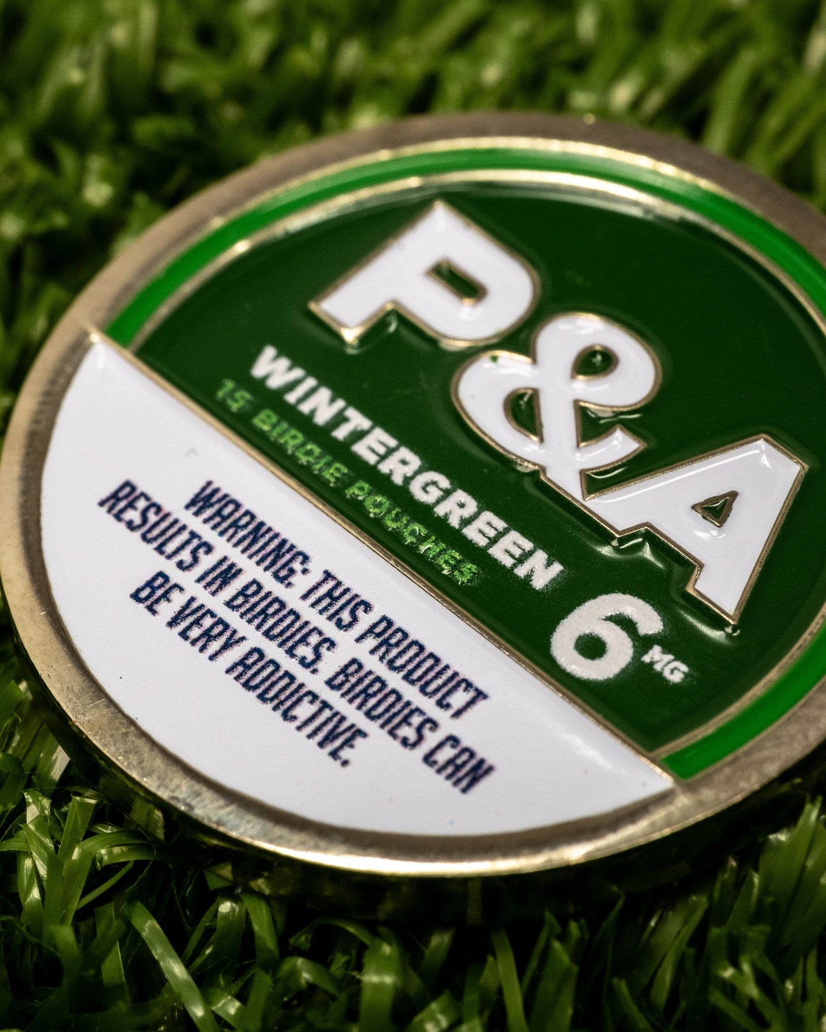 A close-up of a round green and white Pins and Aces 6 Milly - Wintergreen Ball Marker sits on artificial grass next to an enamel golf marker, labeled: "This product results in birdies. Birdies can be very addictive.