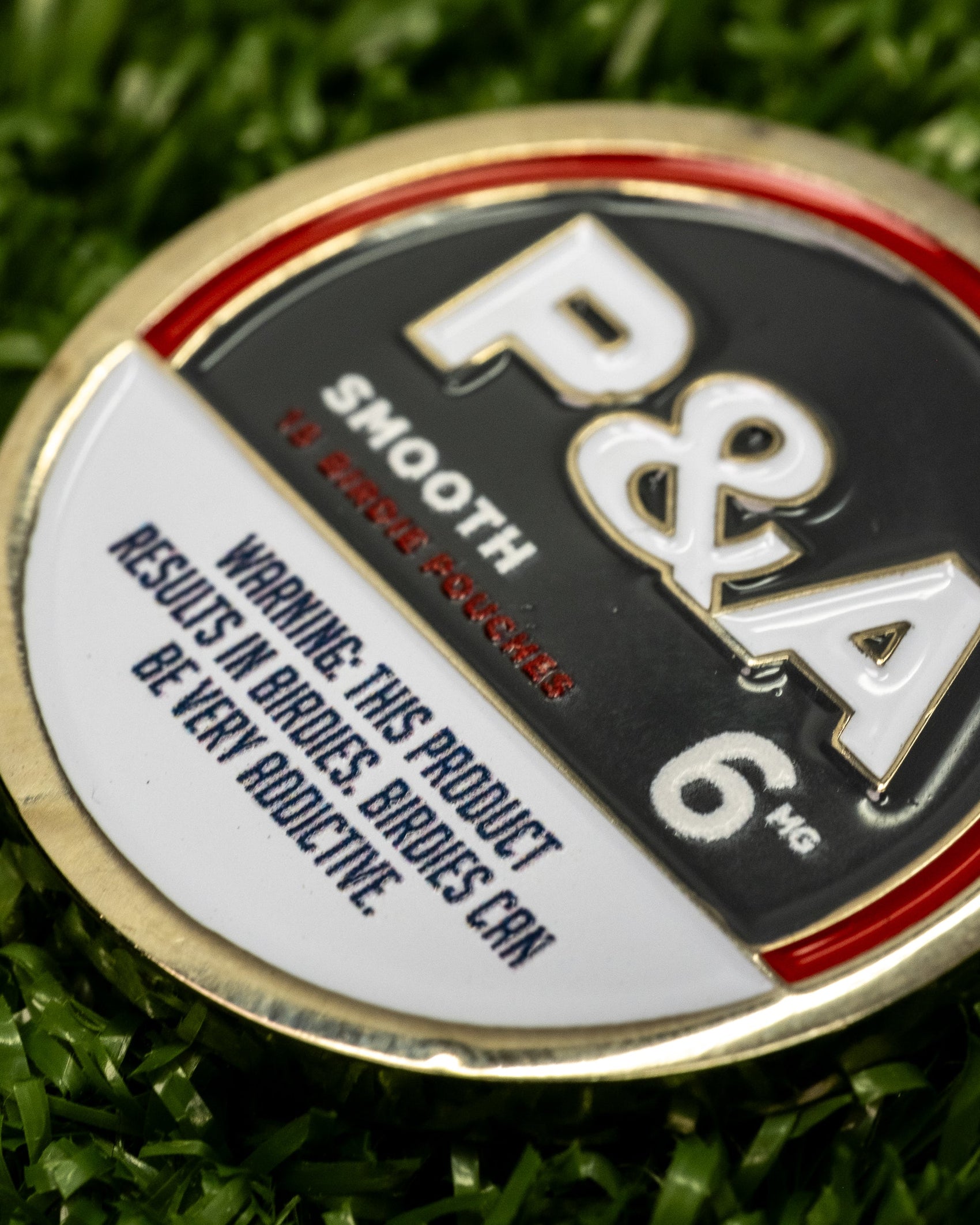 A close-up of the Pins and Aces 6 Milly - Smooth Ball Marker on green grass, built for magnetic surfaces, and featuring a warning: This product results in birdies. Birdies can be very addictive.