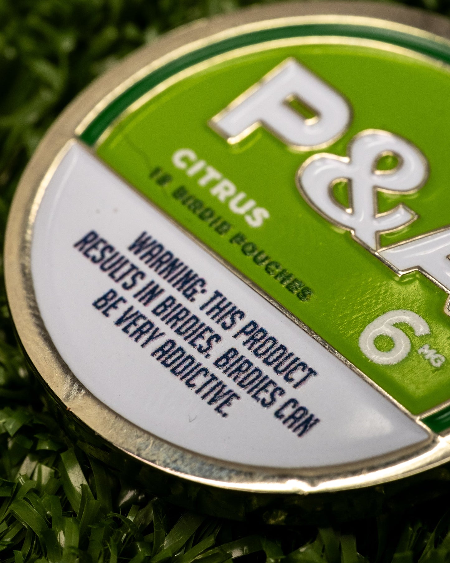 A close-up of a round green and white tin labeled "Pins and Aces 6 Milly - Citrus Ball Marker." The enamel-painted tin rests on grass, warning: THIS PRODUCT RESULTS IN BIRDIES. BIRDIES CAN BE VERY ADDICTIVE.