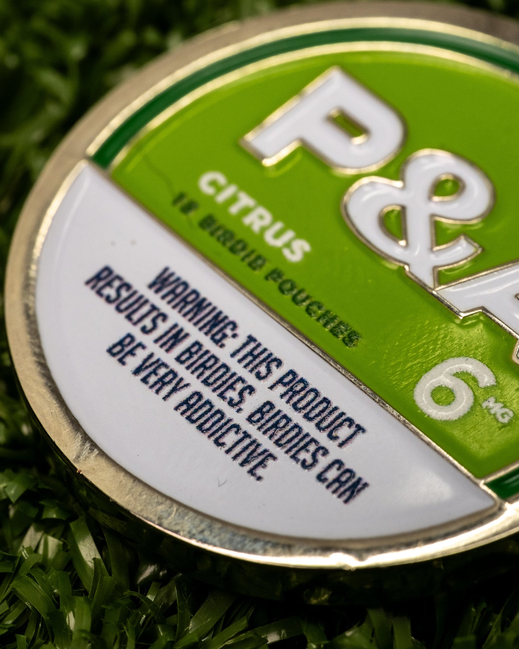 A close-up of a round green and white tin labeled "Pins and Aces 6 Milly - Citrus Ball Marker." The enamel-painted tin rests on grass, warning: THIS PRODUCT RESULTS IN BIRDIES. BIRDIES CAN BE VERY ADDICTIVE.