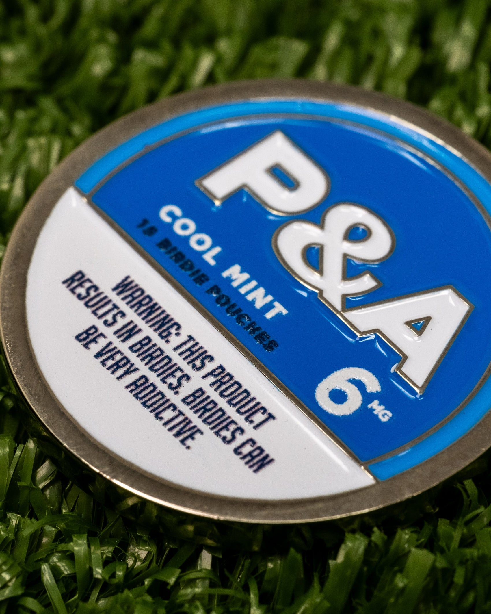 A round blue and silver tin labeled "6 Milly - Cool Mint Ball Marker" by Pins and Aces rests on artificial grass, its magnetic lid perfect for ball markers. Warning: This product results in birdies. Birdies can be very addictive.