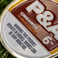 Close-up of a round tin labeled 6 Milly - Cinnamon Ball Marker by Pins and Aces, along with an enamel golf marker, on green grass. Warning: This product results in burns. Burns can be very addictive.