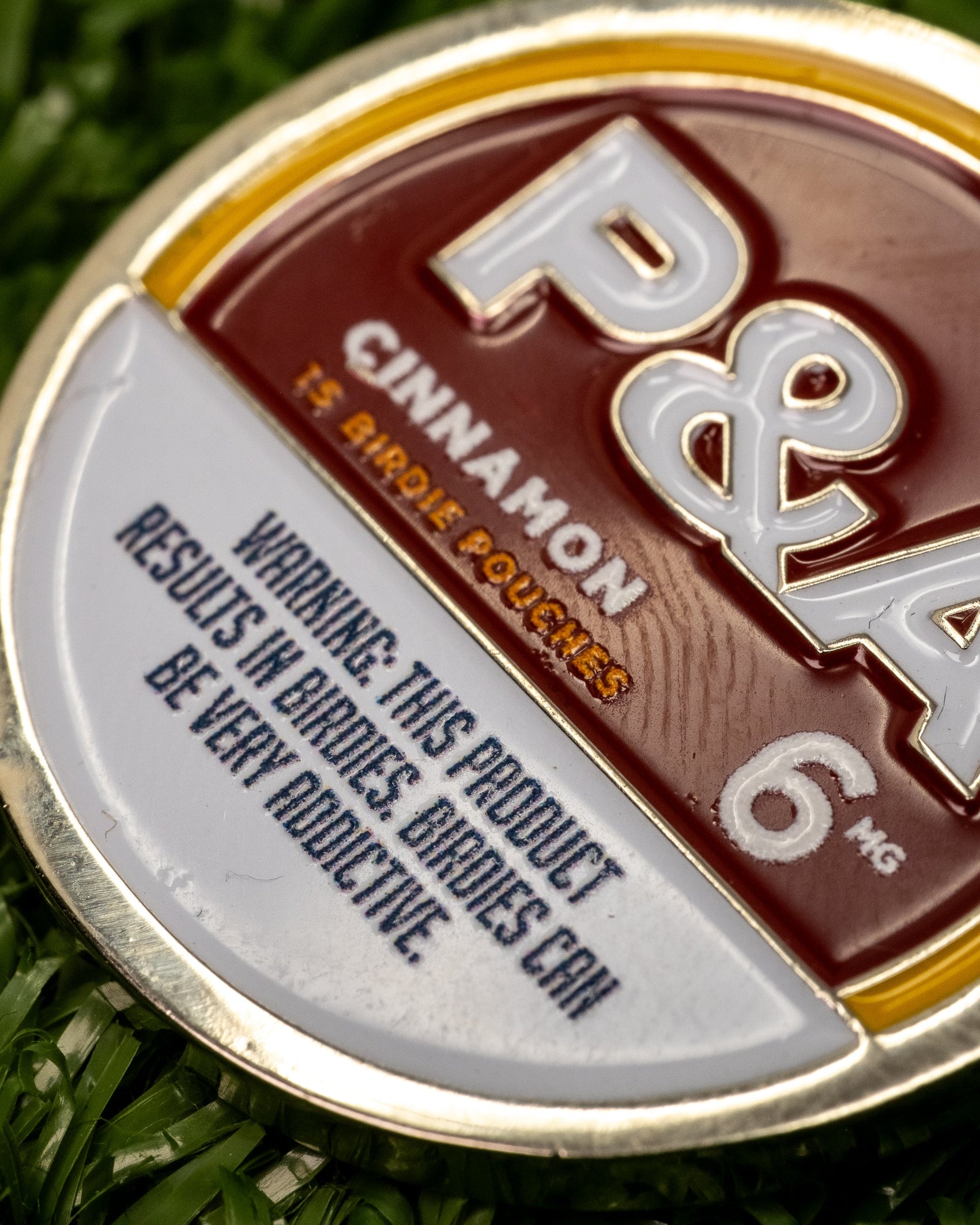 Close-up of a round tin labeled 6 Milly - Cinnamon Ball Marker by Pins and Aces, along with an enamel golf marker, on green grass. Warning: This product results in burns. Burns can be very addictive.