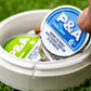A hand picks up a blue 6 Milly - Cool Mint Ball Marker tin by Pins and Aces from a white container with another green-labeled tin, on artificial grass. The magnetic surface is popular for ball markers and the tin warns it may be addictive.
