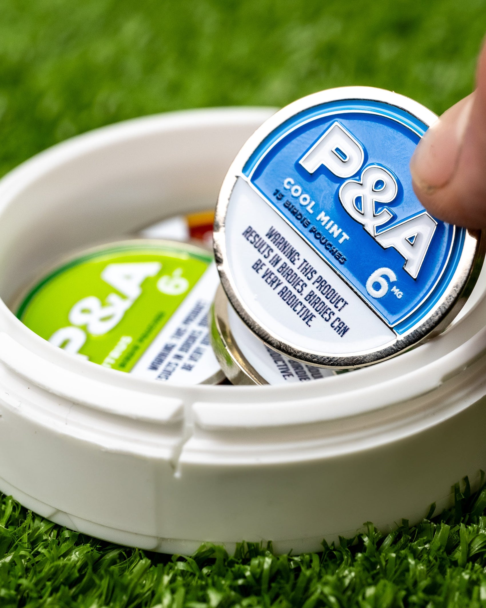 A hand picks up a blue 6 Milly - Cool Mint Ball Marker tin by Pins and Aces from a white container with another green-labeled tin, on artificial grass. The magnetic surface is popular for ball markers and the tin warns it may be addictive.