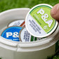 A hand lifts a green-labeled 6 Milly - Citrus Ball Marker tin from an open white container on green turf. The container holds other Pins and Aces tins, such as Cool Mint, each enamel-coated and designed to store golf ball markers.
