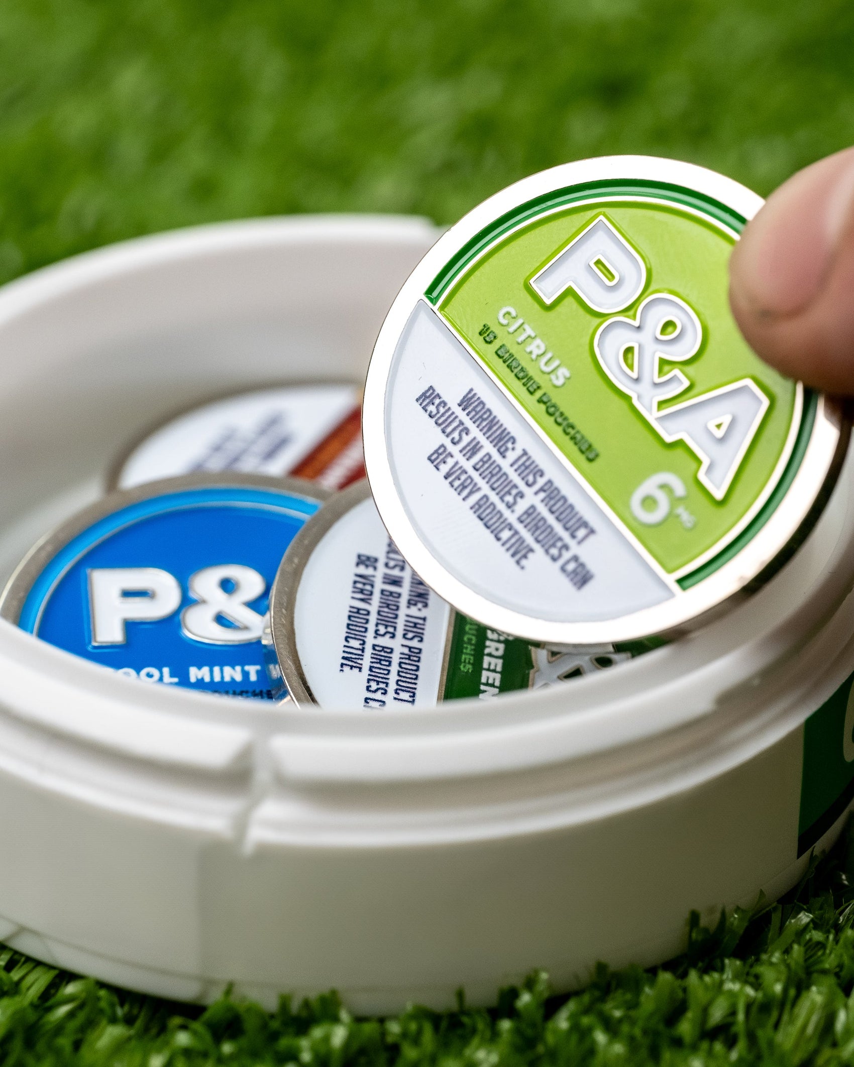 A hand lifts a green-labeled 6 Milly - Citrus Ball Marker tin from an open white container on green turf. The container holds other Pins and Aces tins, such as Cool Mint, each enamel-coated and designed to store golf ball markers.