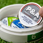 A hand lifts a round 6 Milly - Smooth Ball Marker by Pins and Aces, with a nicotine warning, from a white container of similar tins. The container rests on artificial grass on a magnetic ball marker surface.