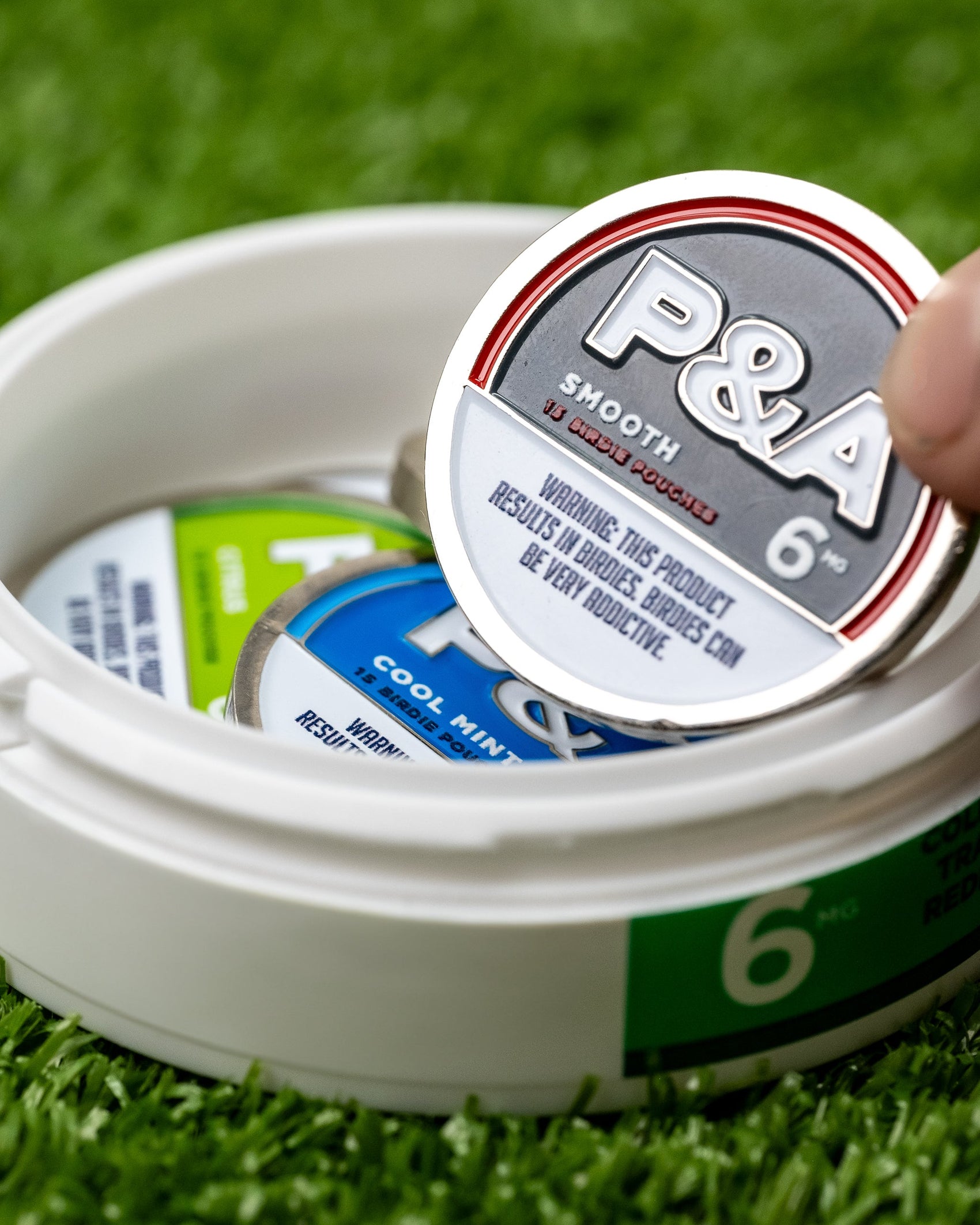 A hand lifts a round 6 Milly - Smooth Ball Marker by Pins and Aces, with a nicotine warning, from a white container of similar tins. The container rests on artificial grass on a magnetic ball marker surface.