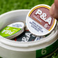A hand picks up a round tin labeled “6 Milly - Cinnamon Ball Marker” by Pins and Aces from an open white container holding similar tins and a shiny enamel golf marker, all set on green artificial grass.