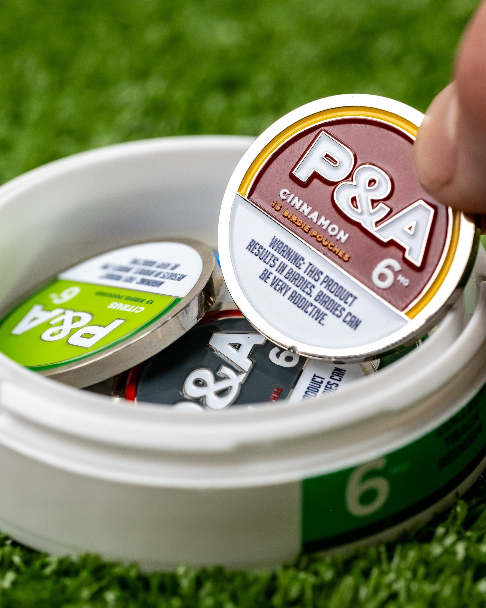 A hand picks up a round tin labeled “6 Milly - Cinnamon Ball Marker” by Pins and Aces from an open white container holding similar tins and a shiny enamel golf marker, all set on green artificial grass.