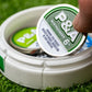 A hand lifts a Pins and Aces 6 Milly - Wintergreen Ball Marker tin from a white case holding another tin and an enamel golf marker. The container sits on green turf, ideal for keeping any golfer’s essentials organized.