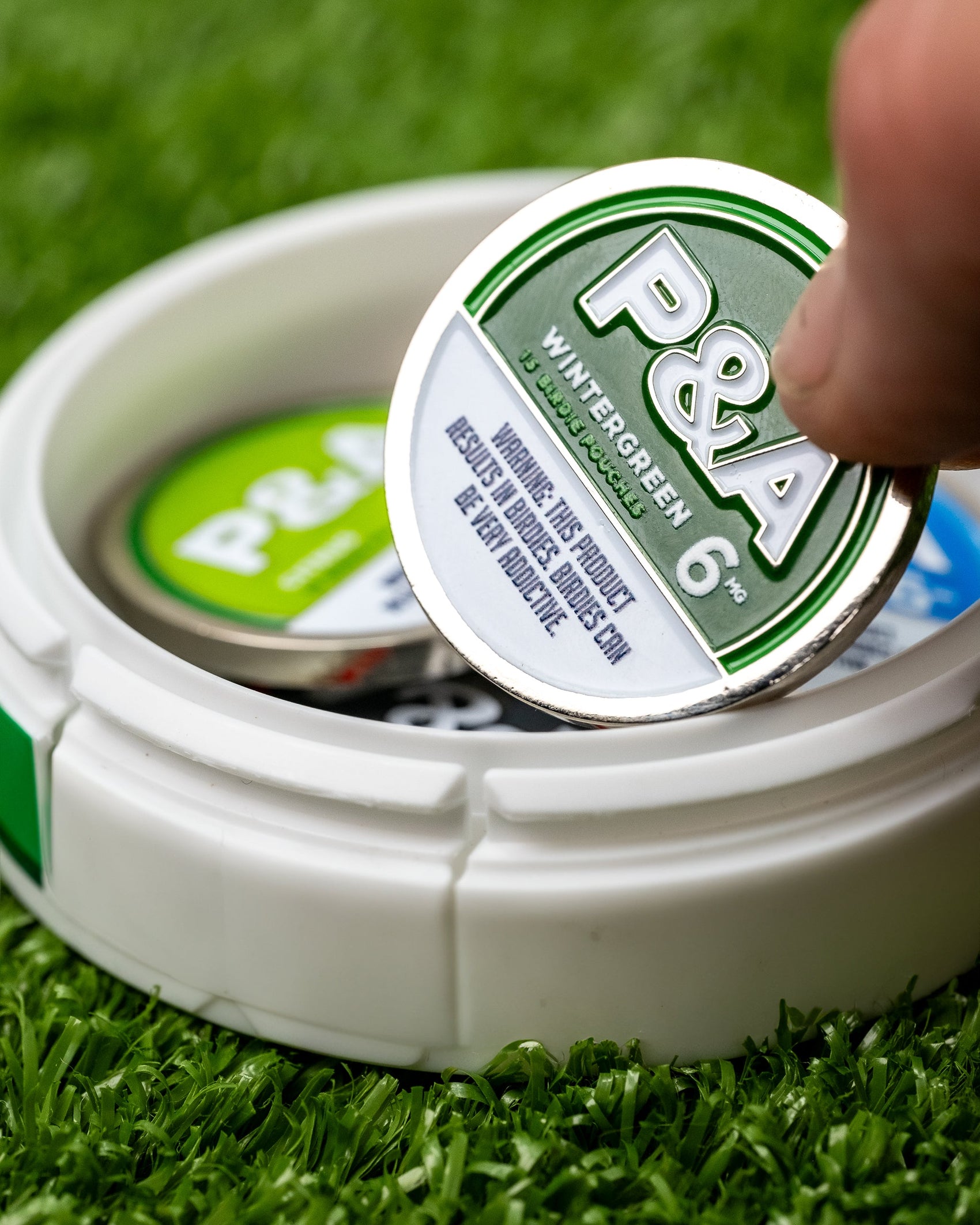 A hand lifts a Pins and Aces 6 Milly - Wintergreen Ball Marker tin from a white case holding another tin and an enamel golf marker. The container sits on green turf, ideal for keeping any golfer’s essentials organized.