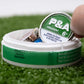 A hand lifts a round can labeled "Pins and Aces 6 Milly - Spearmint Ball Marker" from a white container on artificial green grass, resembling enamel golf markers and displaying an addictiveness warning.