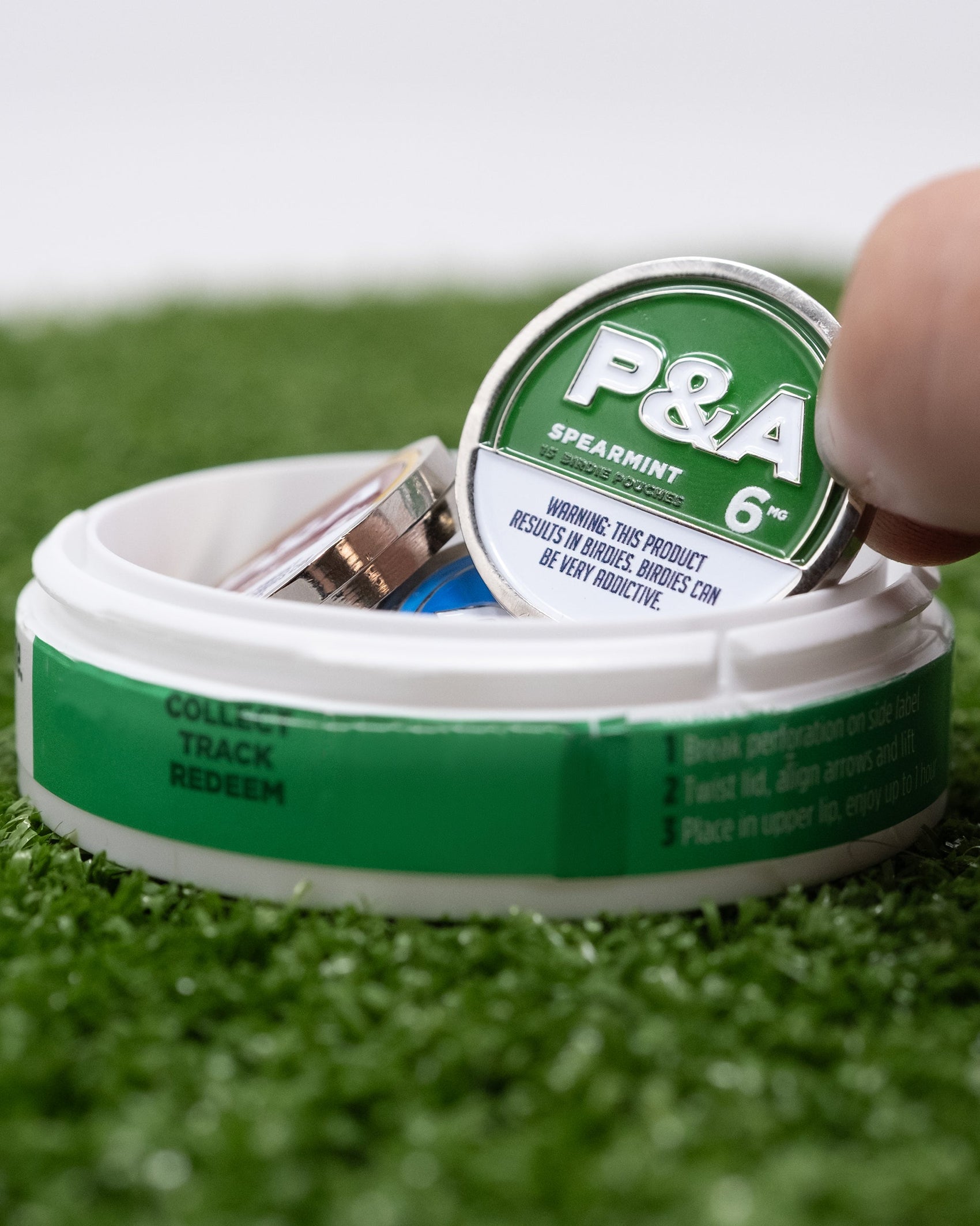 A hand lifts a round can labeled "Pins and Aces 6 Milly - Spearmint Ball Marker" from a white container on artificial green grass, resembling enamel golf markers and displaying an addictiveness warning.