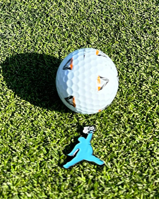 A ChunkMan Ball Marker by Grunk Dolfer rests on green grass, featuring black and orange details. Below is a blue silhouette keychain mid-jump with a golf ball—an eye-catching accessory and unique golf gift.