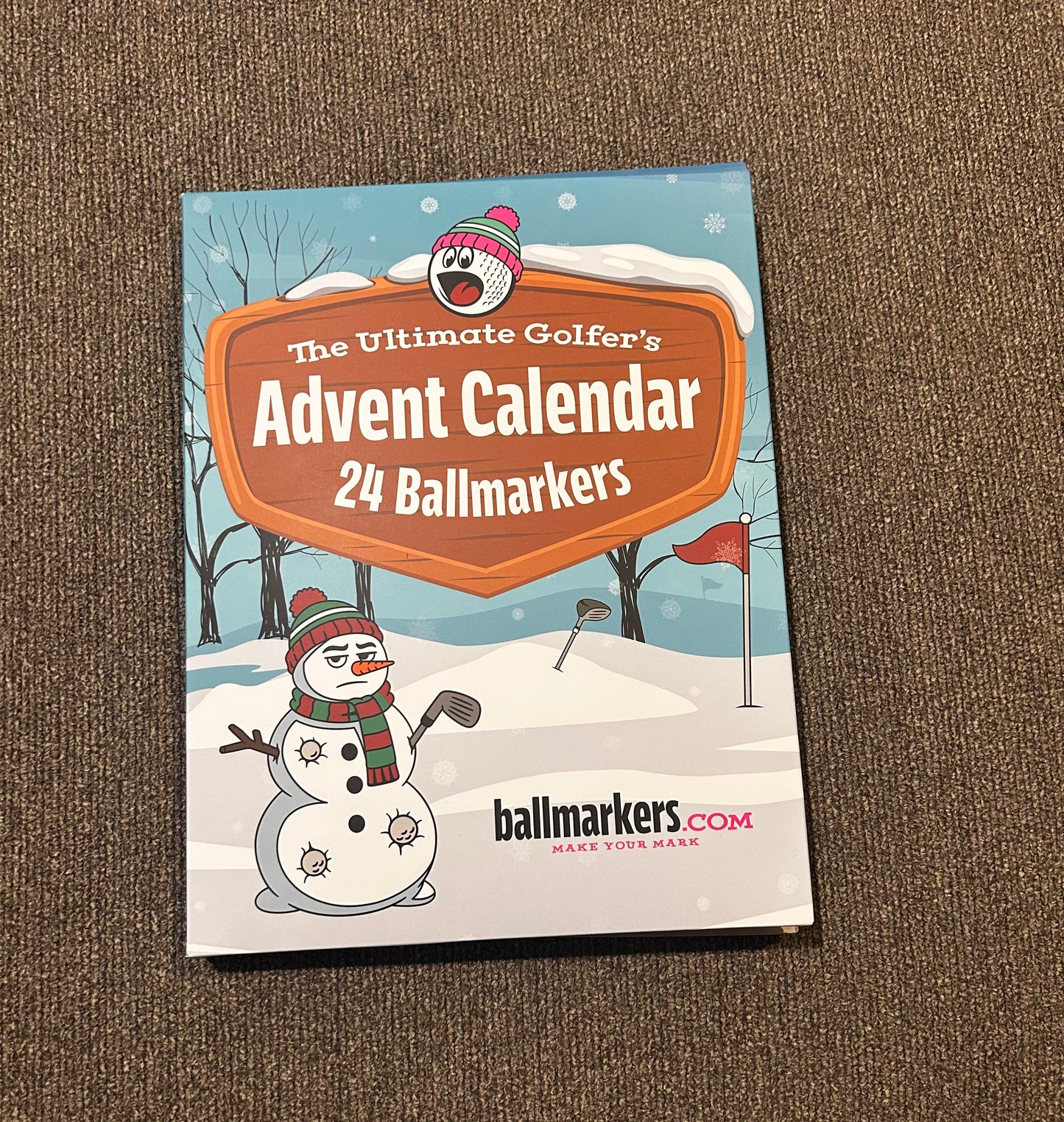 A box labeled The Ultimate Golfers Ballmarkers Advent Calendar by Custom, decorated with snowman and golf-themed winter illustrations, is placed on a brown carpet.