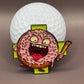 The Donut A Hole Ball Marker by Crown Golf Designs features a pink-frosted donut with cartoon eyes, arms, and legs holding a golf club in front of a white golf ball, with an open mouth and wild look. A green cross appears in the background.