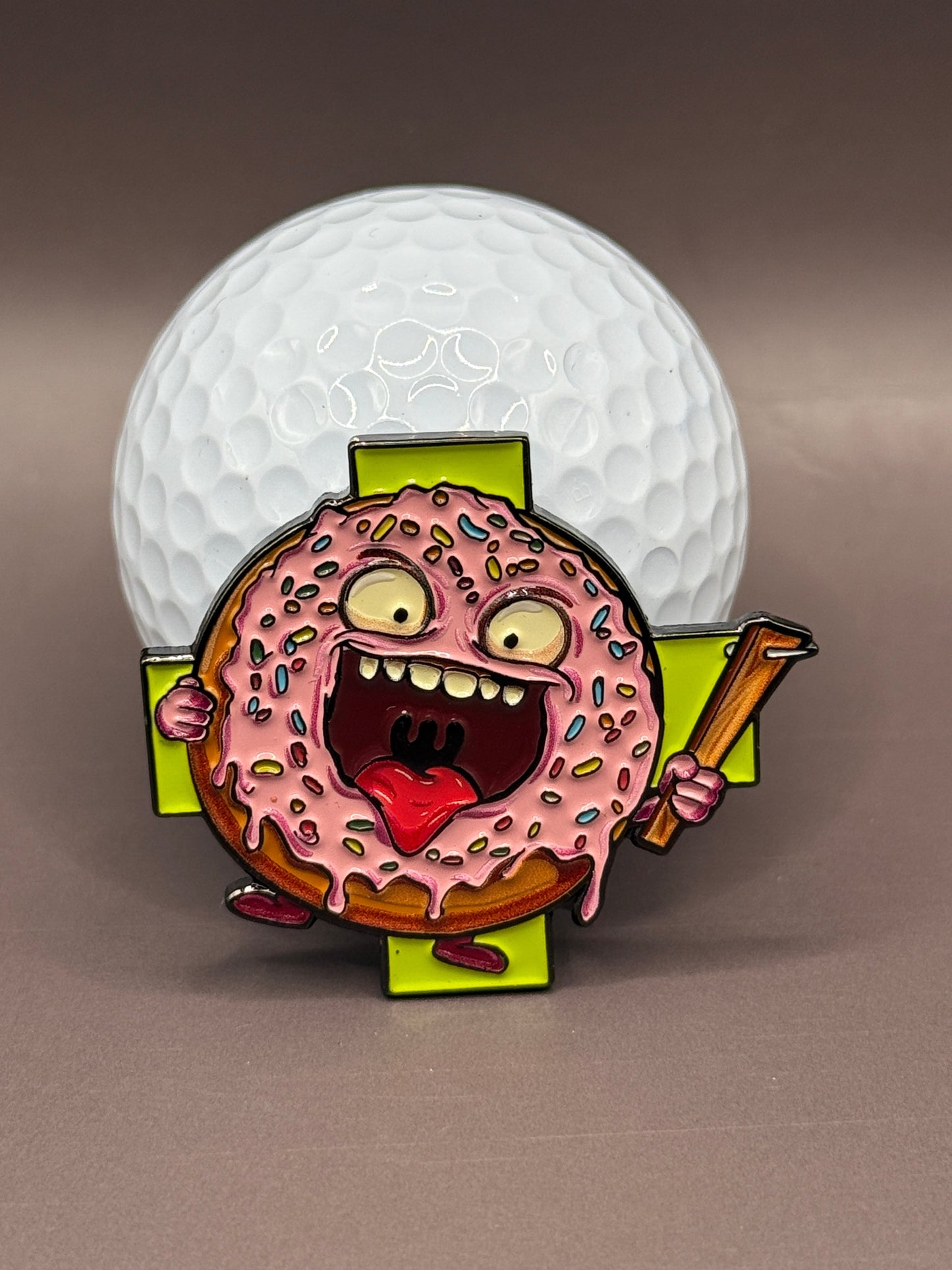 The Donut A Hole Ball Marker by Crown Golf Designs features a pink-frosted donut with cartoon eyes, arms, and legs holding a golf club in front of a white golf ball, with an open mouth and wild look. A green cross appears in the background.