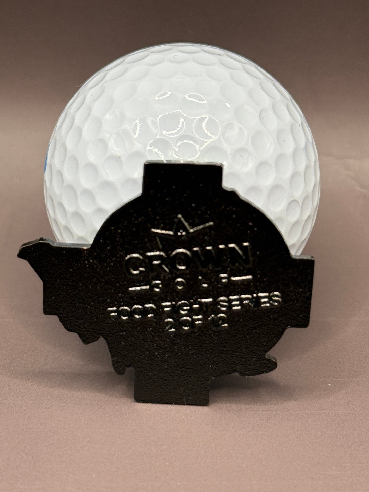 A white golf ball is partially covered by a black Donut A Hole Ball Marker from Crown Golf Designs, engraved with CROWN ROYAL FOOD TOUR SERIES 2014 and a crown logo.