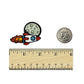 The Kolorspun Rocket Ship Golf Ball Marker is a small enamel pin featuring a rocket and moon, shown next to a U.S. quarter and ruler for scale. Measuring about 1.5 inches, it makes a unique golf gift or custom ball marker.