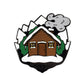 Kolorspun's Log Cabin Golf Ball Marker features a stylized wooden cabin with a brown door, snowy trees, mountains, and a gray cloud above the snow-covered roof—an ideal custom golf gift or unique ball marker.