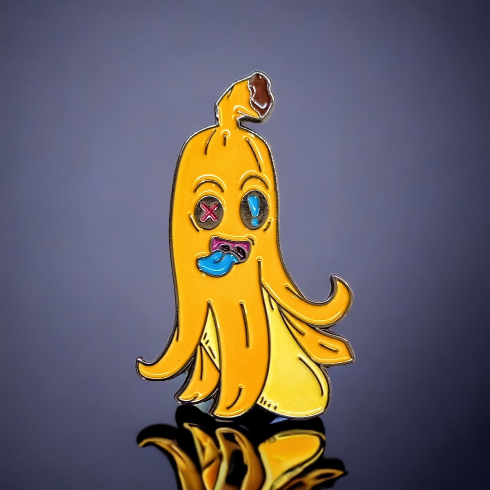 The Peely Ball Marker by Crown Golf Designs is a colorful enamel pin shaped like a cartoon banana peel with a goofy face, X-shaped eye, blue tongue, and brown spot—an eye-catching golf accessory against a dark reflective surface.