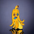 The Peely Ball Marker by Crown Golf Designs is a colorful enamel pin shaped like a cartoon banana peel with a goofy face, X-shaped eye, blue tongue, and brown spot—an eye-catching golf accessory against a dark reflective surface.