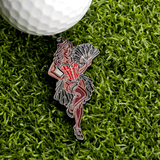 A golf ball on artificial grass sits beside the Burlesque Girl Ball Marker from Ballmarkers.com, featuring a playful woman in a red corset, black skirt, and top hat holding a fan.