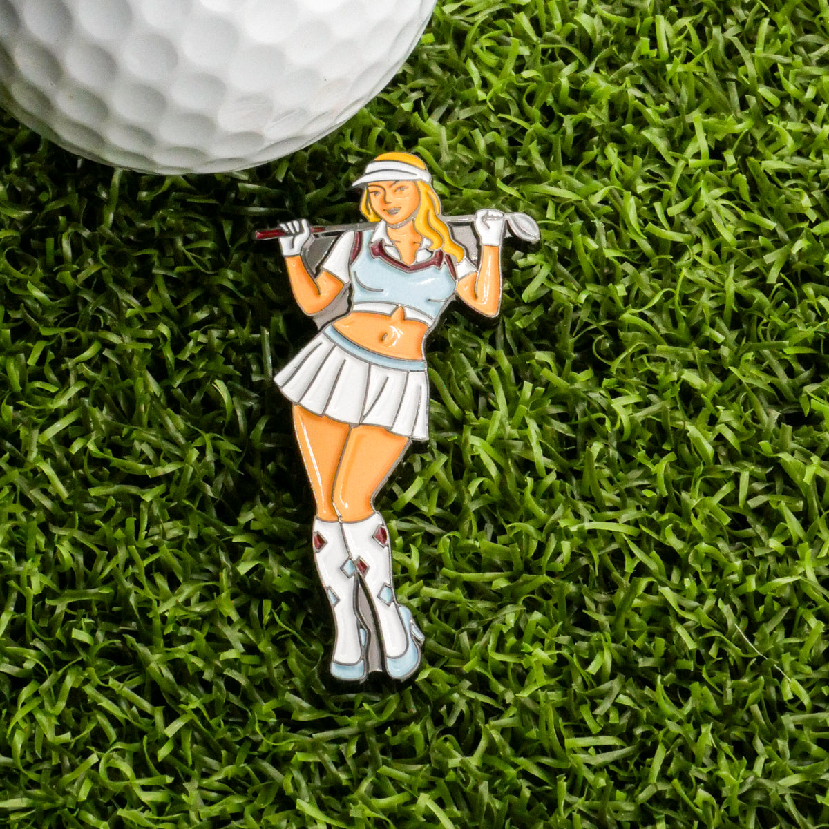 A golf ball on artificial grass beside the Golfer Girl Ball Marker by Ballmarkers.com—shaped like a blonde golfer in a white skirt and blue top—makes a unique golf gift and accessory.