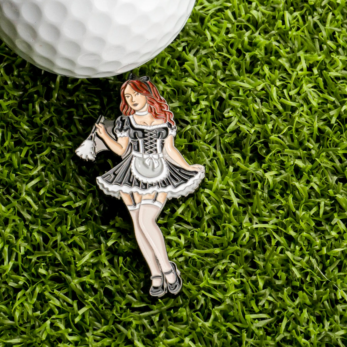A golf ball sits on green grass next to a Sexy Maid Ball Marker from Ballmarkers.com—an enamel pin featuring a maid in black and white with a feather duster—for unique golf accessories.
