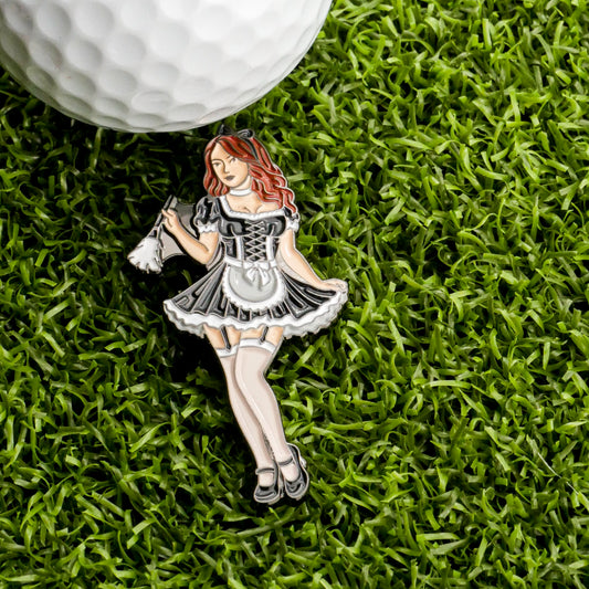 A golf ball sits on green grass next to a Sexy Maid Ball Marker from Ballmarkers.com—an enamel pin featuring a maid in black and white with a feather duster—for unique golf accessories.