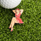 A golf ball sits on artificial grass beside the Pin-up Girl Ball Marker by Ballmarkers.com, depicting a woman in a red dress and heels posing with hands behind her head and legs crossed.