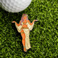 A golf ball on artificial grass alongside the Toga Goddess Ball Marker by Ballmarkers.com, featuring a woman in a flowing dress with outstretched arms, headdress, and sandals.