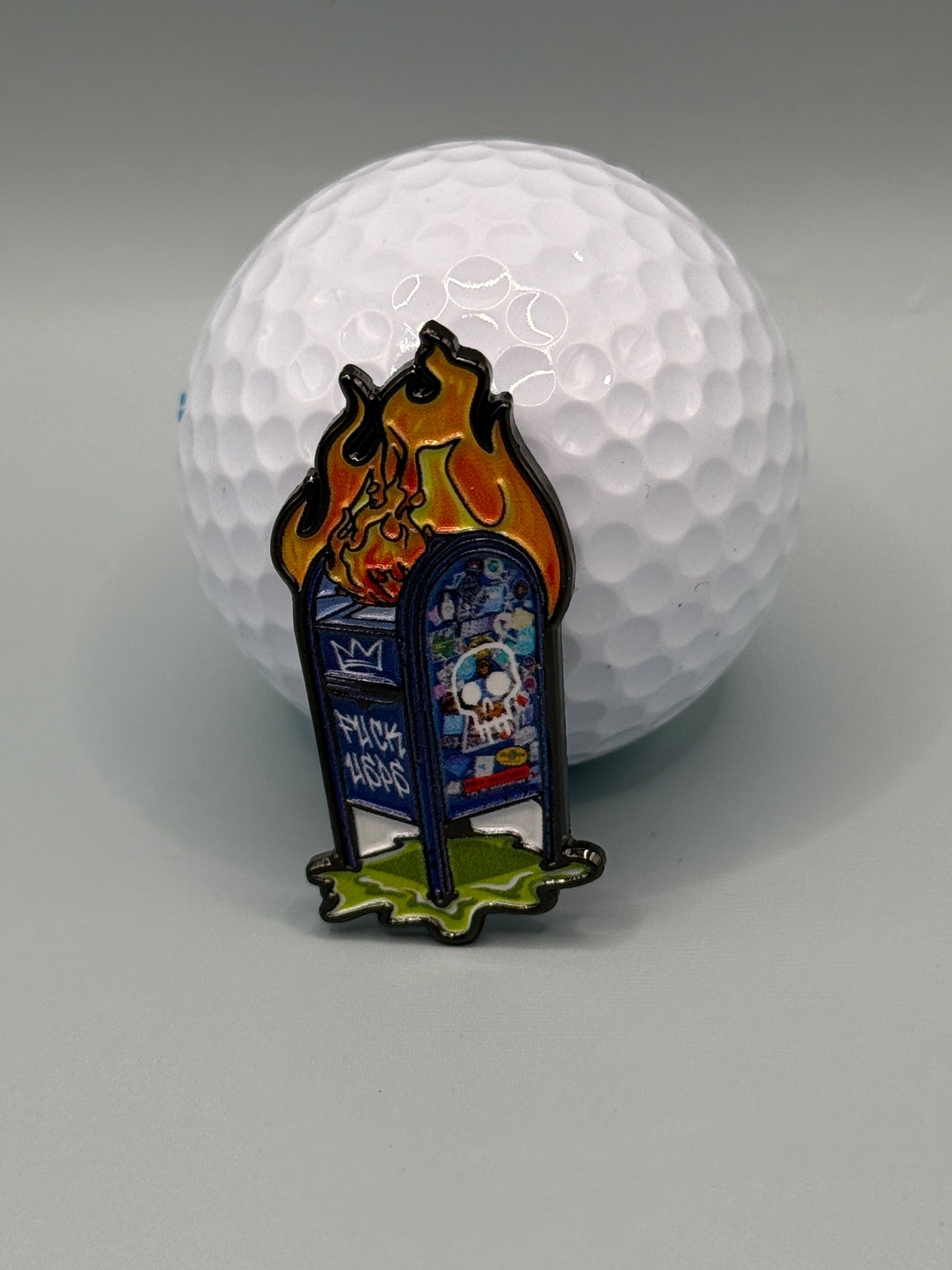 A golf ball sits behind the "UPS Dumpster Fire" enamel pin by Crown Golf Designs, showing a blue mailbox with graffiti and cartoon flames, and featuring a magnetic backing for easy display.
