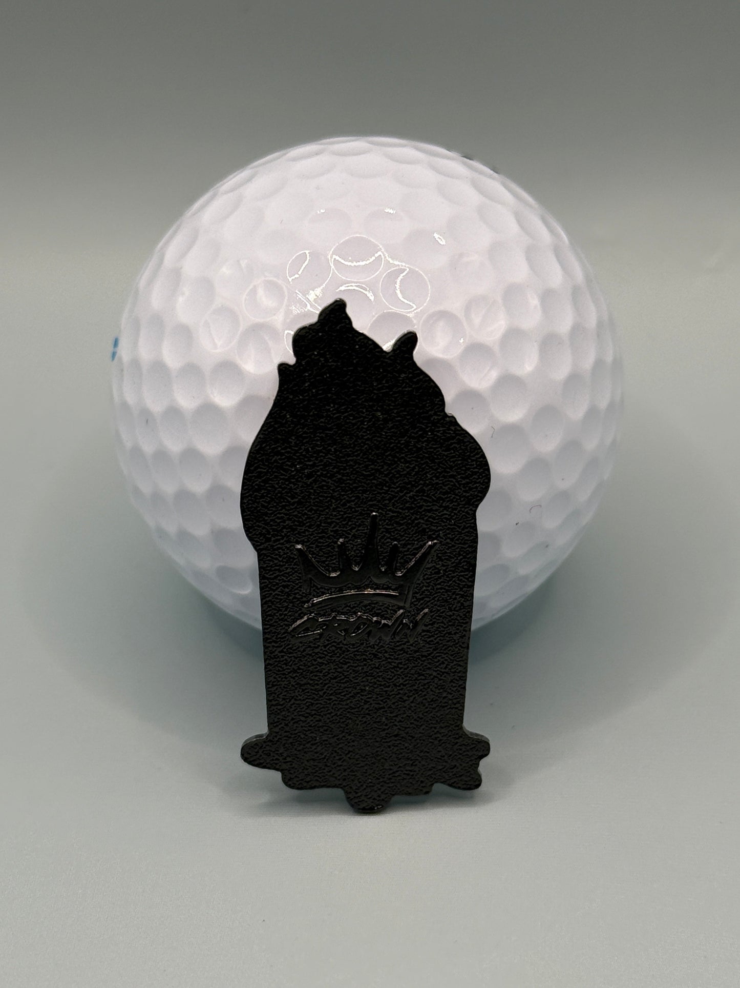 A UPS Dumpster Fire golf ball from Crown Golf Designs sits on a gray surface with a small, magnetic, crown-shaped marker featuring a raised crown design placed in front of it.