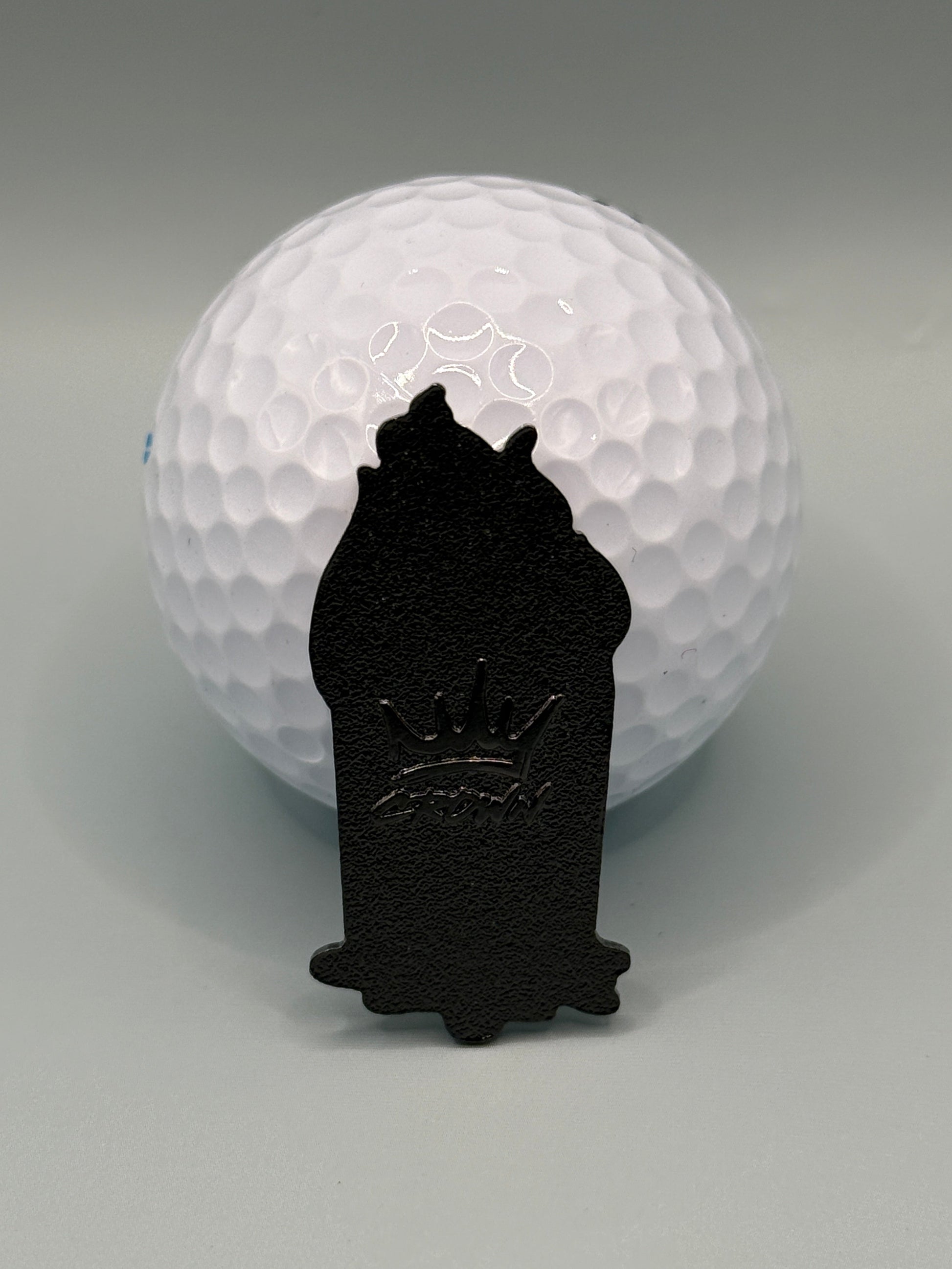 A UPS Dumpster Fire golf ball from Crown Golf Designs sits on a gray surface with a small, magnetic, crown-shaped marker featuring a raised crown design placed in front of it.