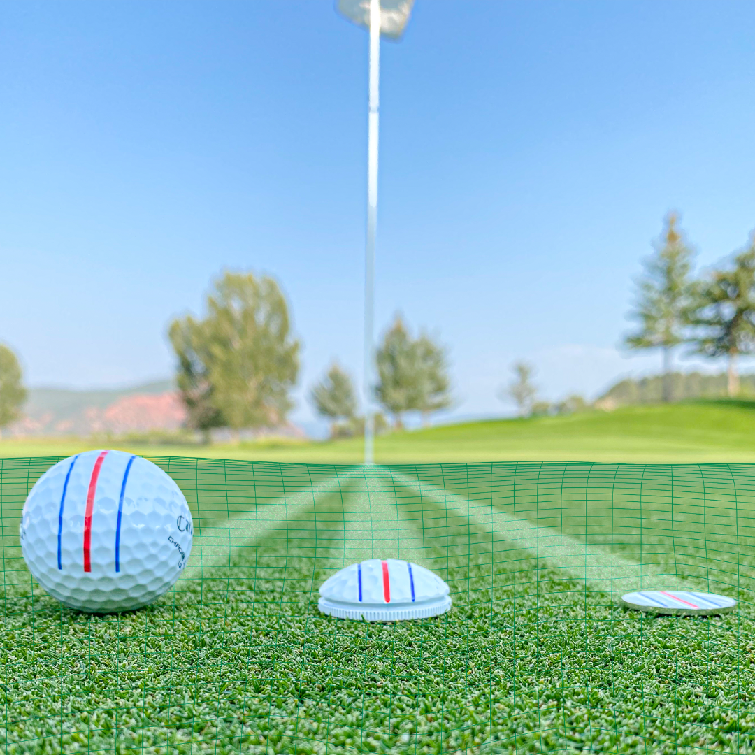 A close-up of the OnPointGolf.us Three Rail Ball Marker, a golf ball, and a half ball on green turf under clear skies, with a flagpole and trees gently blurred in the background.