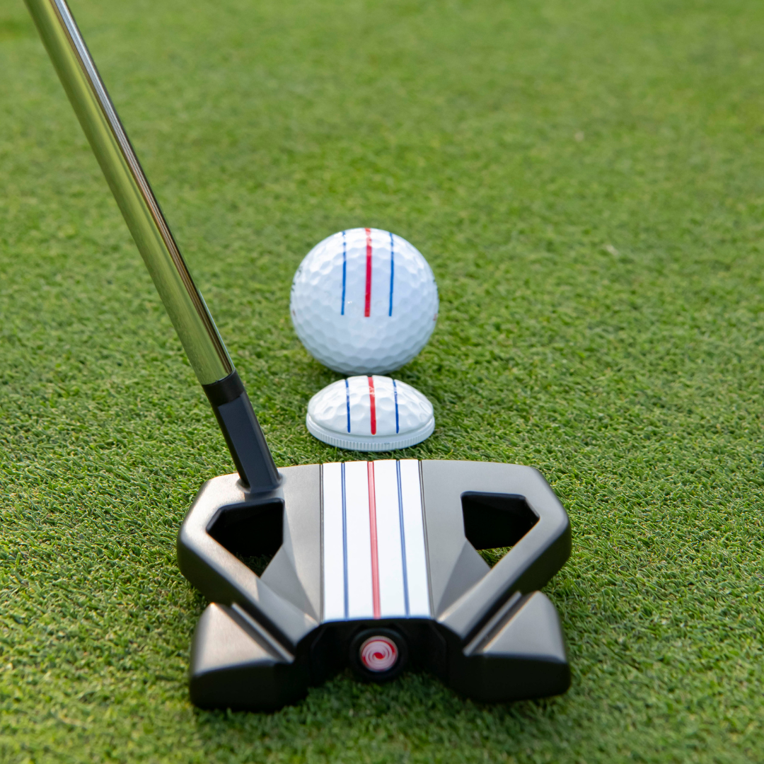 A close-up of the OnPointGolf.us Three Rail Ball Marker and a putter lined up with a golf ball on the green, both displaying red and blue alignment lines to help aim your shot.
