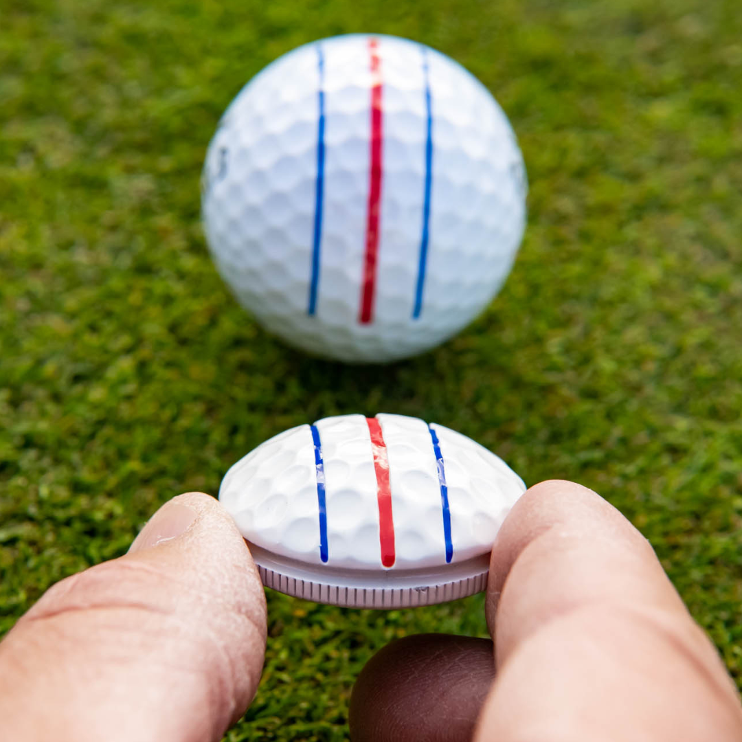 Close-up of two fingers holding the OnPointGolf.us Three Rail Ball Marker with blue and red lines, aligning it with a matching golf ball on green grass.