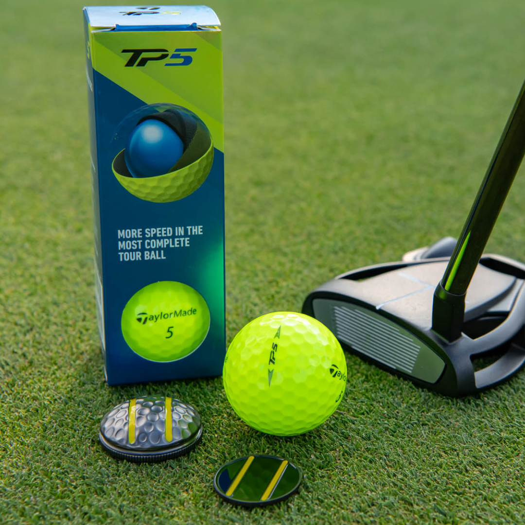 A neon yellow TaylorMade TP5 golf ball, a golf putter, a blue TP5 box, and the OnPointGolf.us Two Rail Ball Marker with extra markers rest on green grass—an ideal putting aid for your next round.