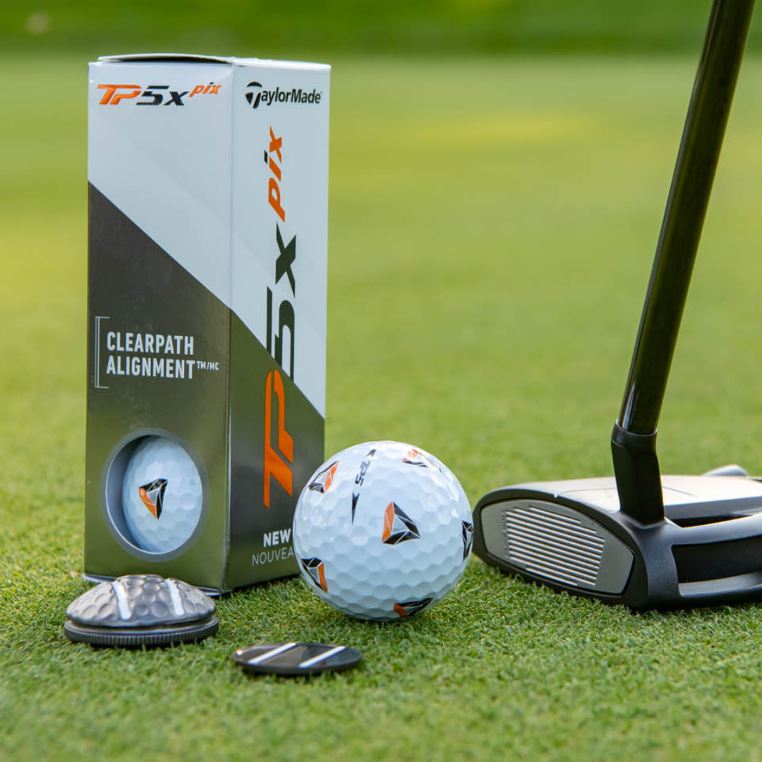A close-up of a TaylorMade TP5x Pix golf ball with orange and black triangles, a putter, and two OnPointGolf.us Two Rail Ball Markers rests on grass next to the golf ball box.