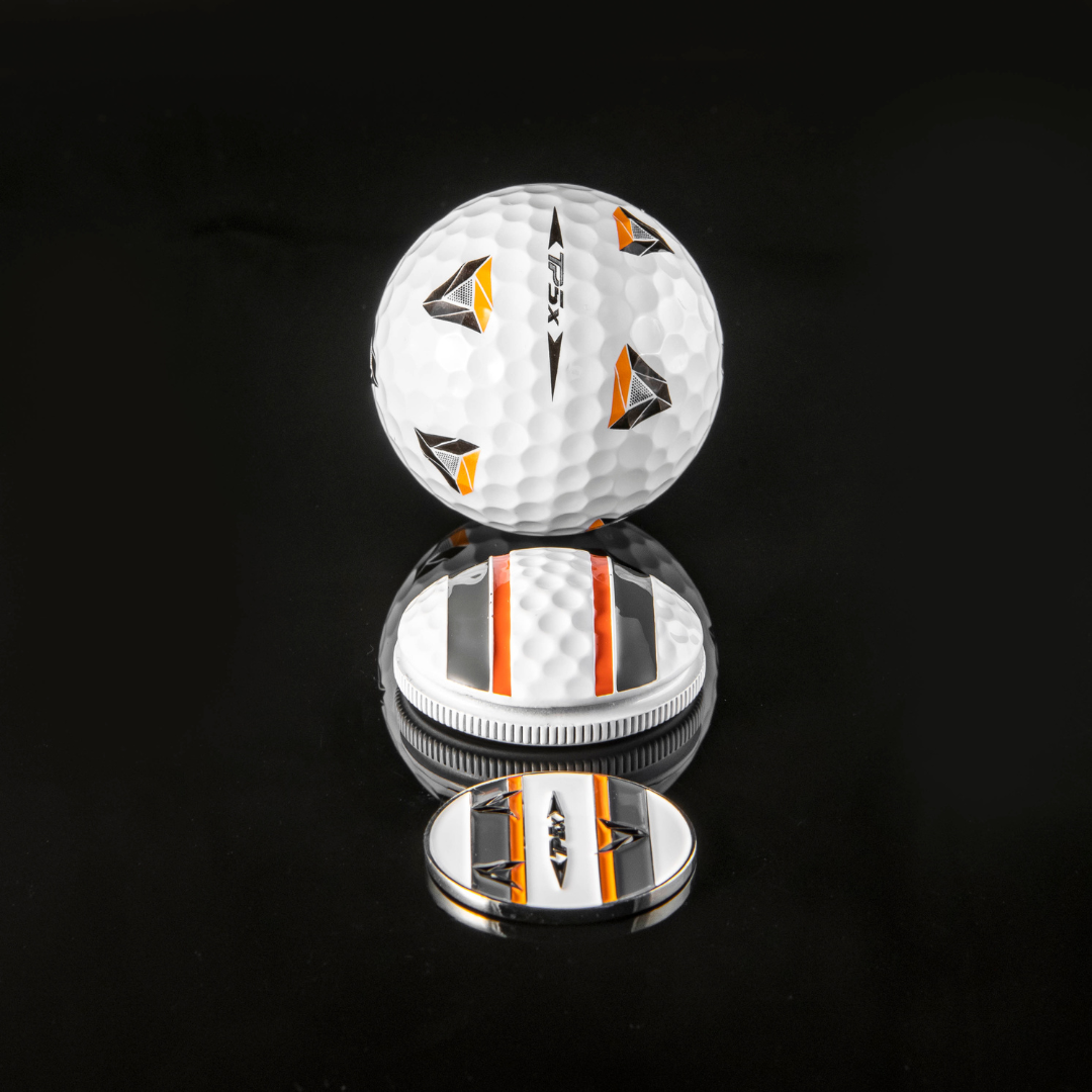 The OnPointGolf.us Two Rail Ball Marker set includes a white golf ball with orange, black, and silver triangular designs, a matching 3D ball marker, and an alignment tool with bold black and orange stripes on a reflective surface.