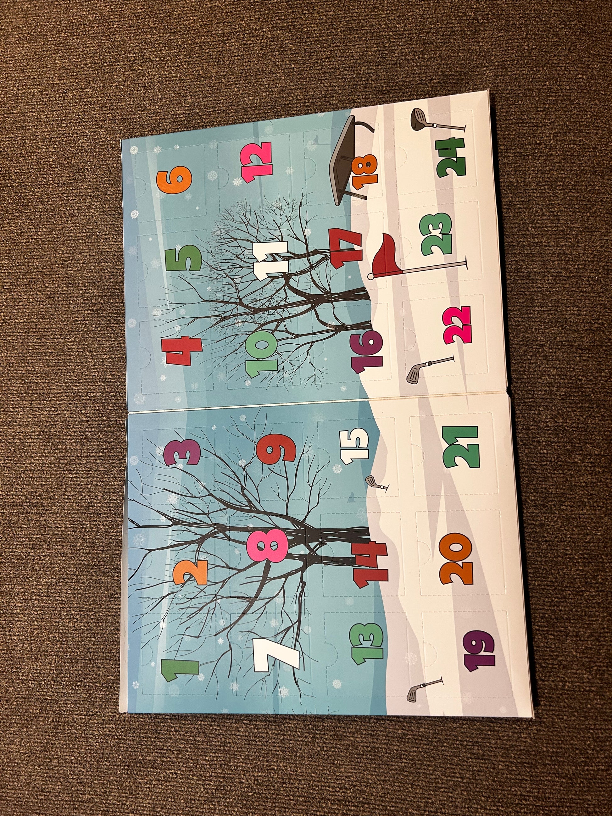 The Ultimate Golfers Ballmarkers Advent Calendar by Custom features colorful, numbered doors from 1 to 21 set against a wintry backdrop with bare trees, snow, and golf-themed elements like clubs and ball markers inspired by Ballmakers.com. The calendar is displayed open on a brown carpet.