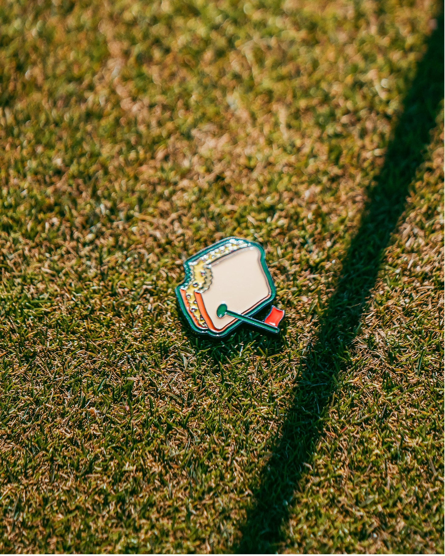 A Pimento Cheese Sandwich Ball Marker by Pins and Aces rests on sunlit grass, casting a long shadow—perfect as a magnetic hat clip or an Augusta-inspired golf marker for your next round.