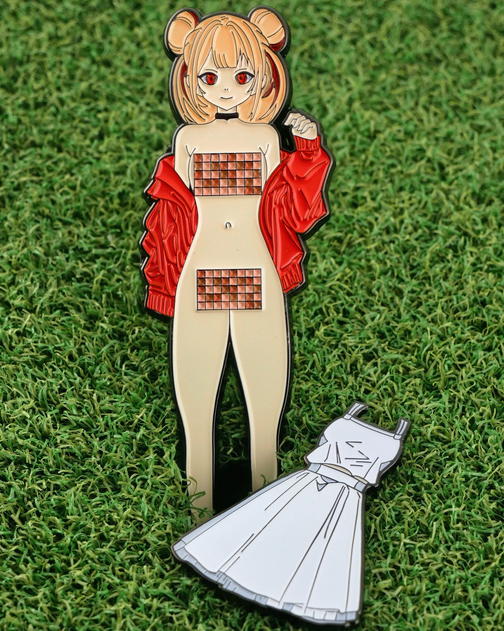 The Japanese Anime golf ball marker from Ballmarkers.com features a girl with bear-ear buns, red jacket, and pixelated bars on grass. A separate white dress pin lies beside her, resembling a divot tool.