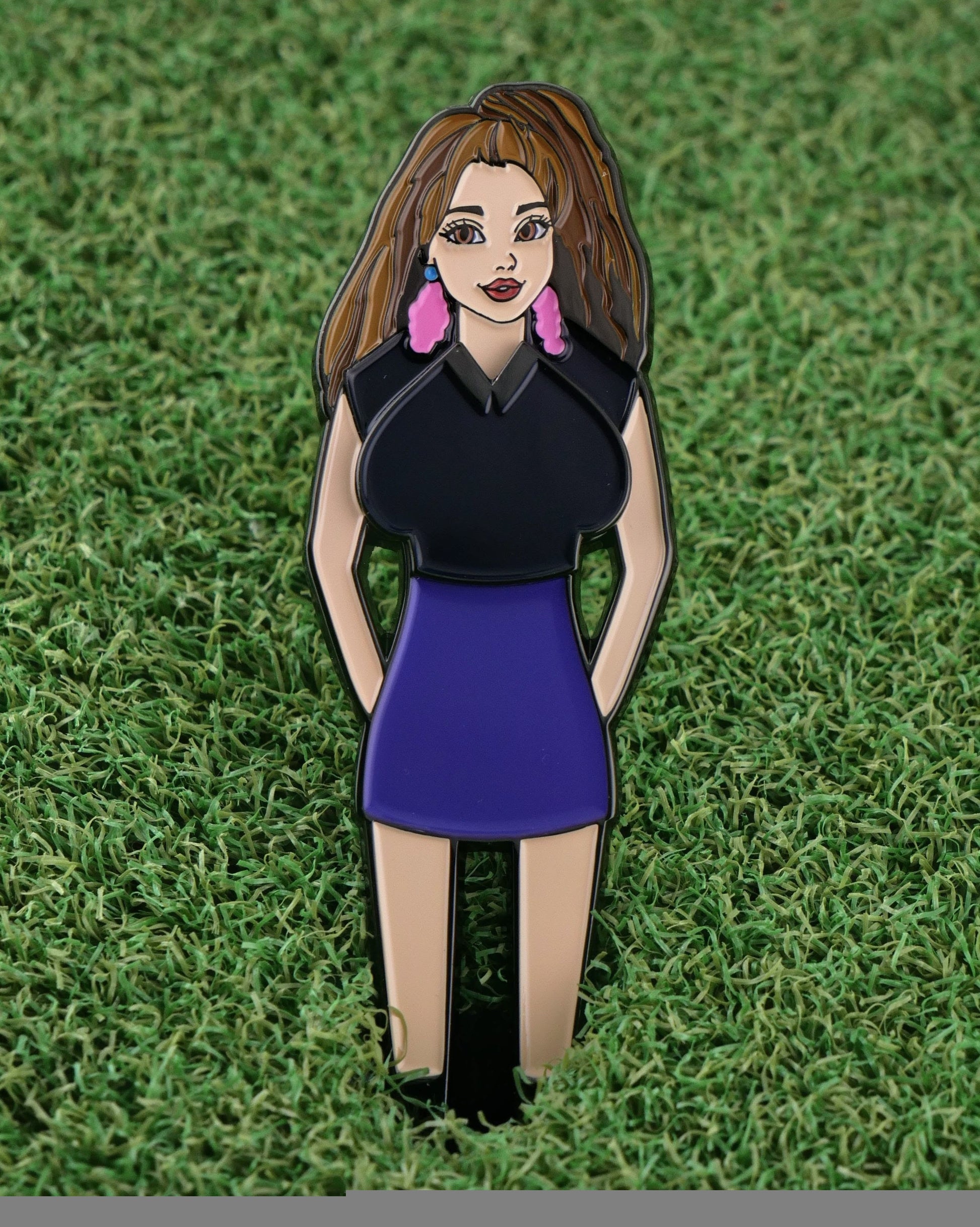 The Trifecta Chick enamel pin by Ballmarkers.com features a woman with long brown hair in a black top and purple skirt on green grass—a unique, funny golf gift that doubles as a golf accessory or ball marker.