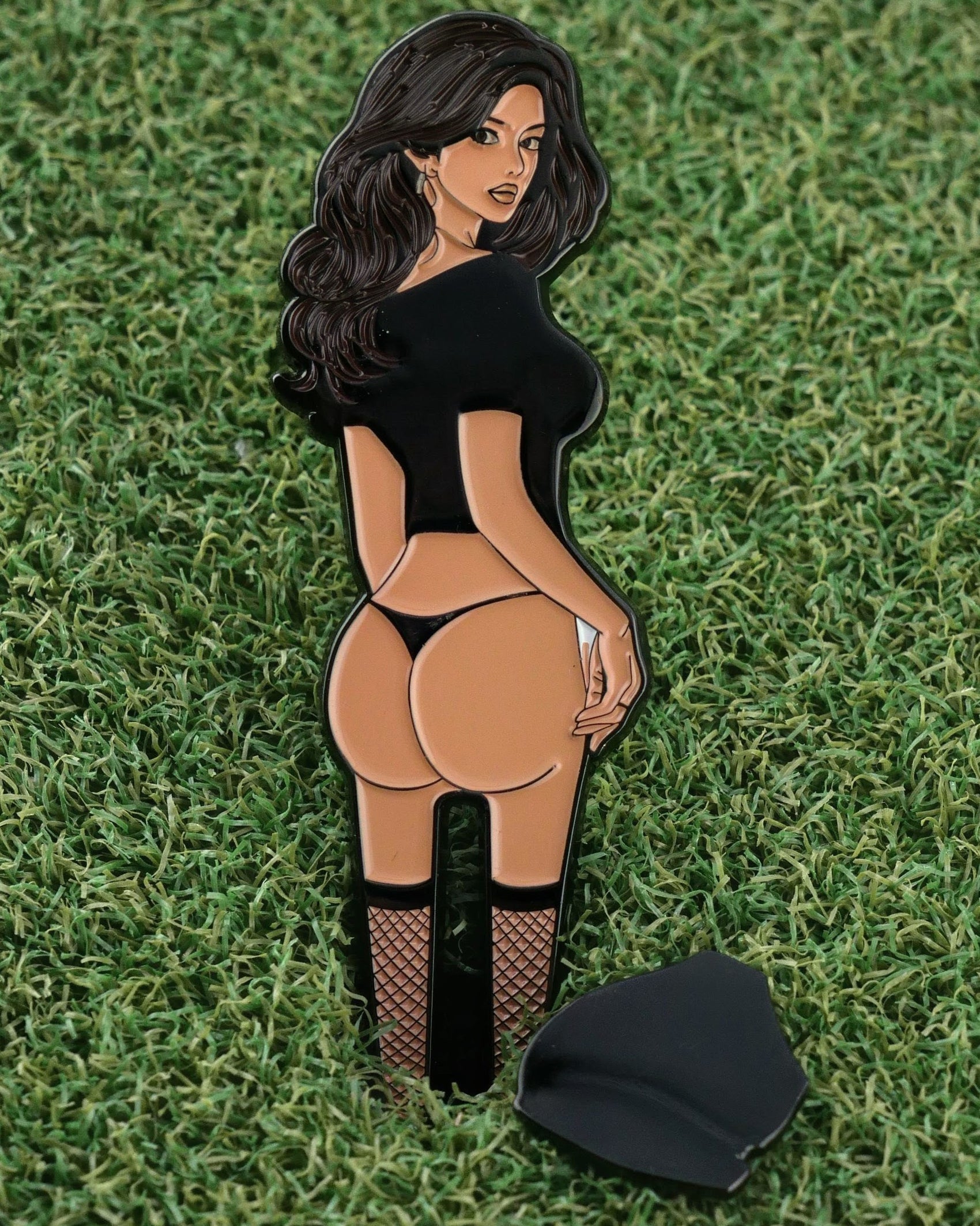 The Thiccc Chick enamel pin by Ballmarkers from Logie Joe’s shows a cartoon woman in a black crop top, thong, and fishnets on grass as a unique golf ball marker. The pin is broken with a detached piece beside it.